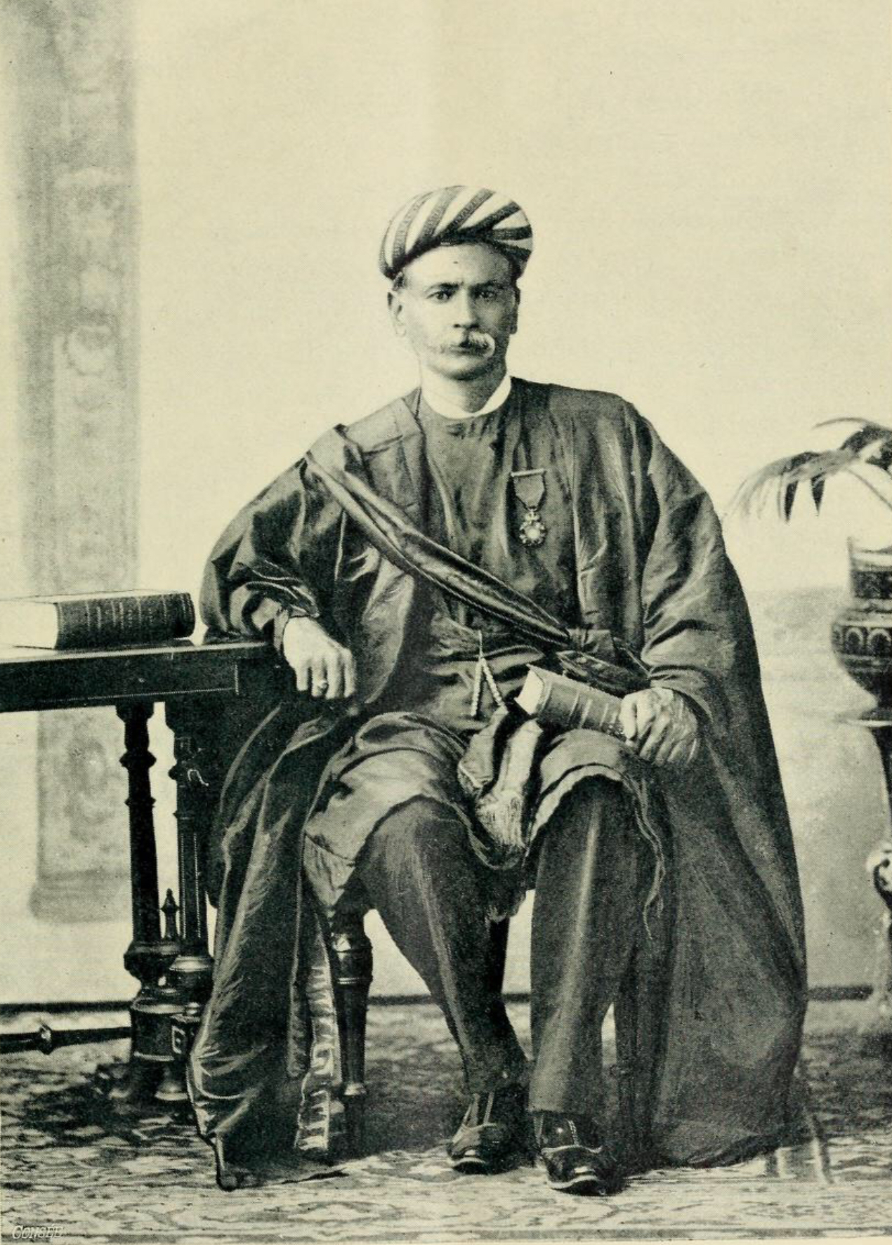 Pratap Chandra Ray seated beside a table with books, wearing a turban and a medal